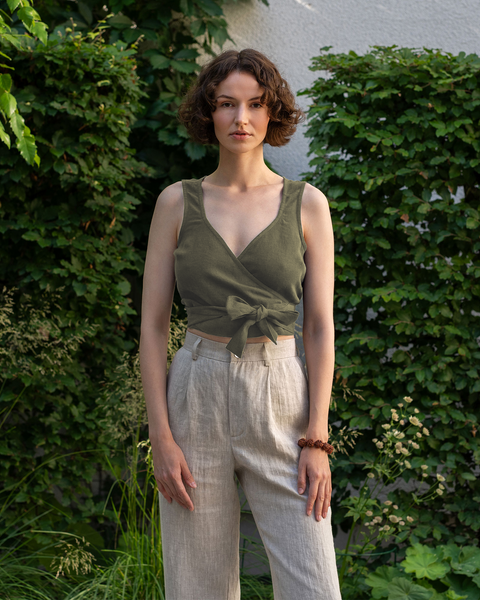 A woman wearing 100% linen sleeveless wrap crop top in the color stone green. She is looking straight and the photo's background is green and full of vegetation.