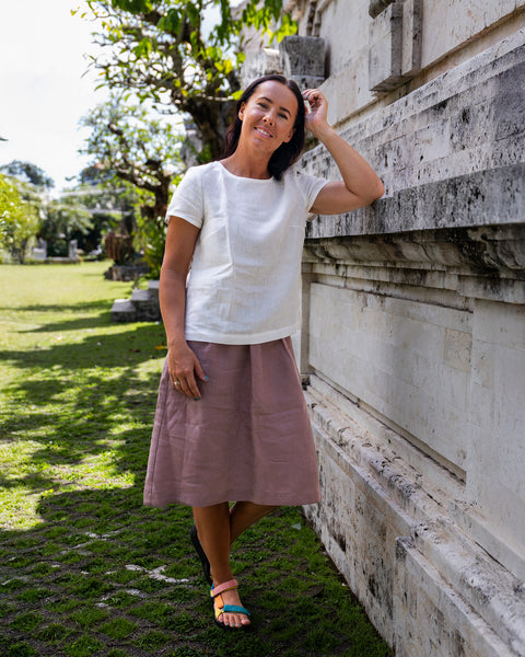 A woman with dark hair smiles while leaning against a white stone wall outdoors. She wears a white short-sleeved top, a dusty pink knee-length skirt, and colourful flat sandals. Her left elbow rests on the wall, and her left hand is near her hair. The background shows a grassy area and trees under a sunny sky.
