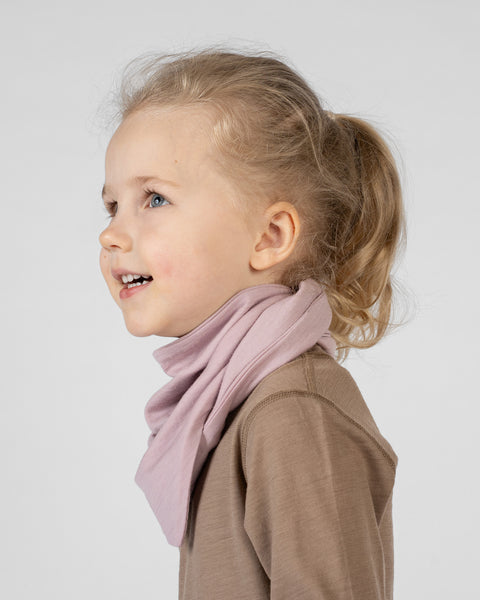 A studio portrait of a young blonde girl looking upwards and smiling slightly. She is seen in profile from the shoulders up. She is wearing a dusty pink bandana tied around her neck, over a light brown long-sleeved shirt. The background is a plain light grey.