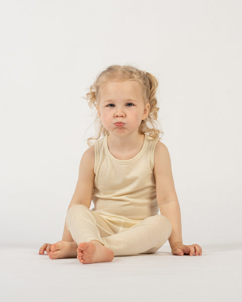 A young child with light curly hair in pigtails is sitting on a white surface against a plain white background. The child is wearing a natural colored sleeveless top and matching leggings. They are looking directly at the camera with a pouty expression, and their bare feet are visible.