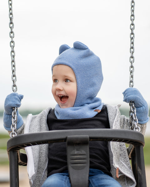 A young child in a light blue menique balaclava with ear-like details, blue gloves, a gray zippered hoodie, and blue jeans, is sitting in a swing. The child has an open mouth and is looking upwards and to their right, seemingly laughing or excited. The background is a blurred outdoor scene with a body of water and distant land.