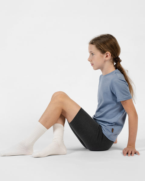 A side-profile view of a young girl sitting on a white surface with her knees bent and leaning back on her hands, modeling charcoal merino cycling shorts, natural ribbed crew socks, and a dusty blue short-sleeve t-shirt.
