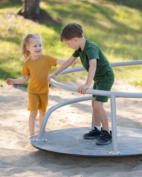 Two children wearing matching Merino wool T-shirts and shorts in yellow and dark green. The lightweight, breathable kids' outfits are showcased in a sunny park setting, perfect for everyday play, family outings, and warm-weather adventures.