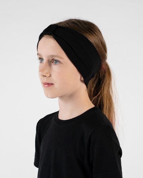 A studio portrait of a young girl looking off to her left in a three-quarter profile against a white background. She is wearing a black short-sleeved t-shirt and a black ribbon headband with a knot detail on top.