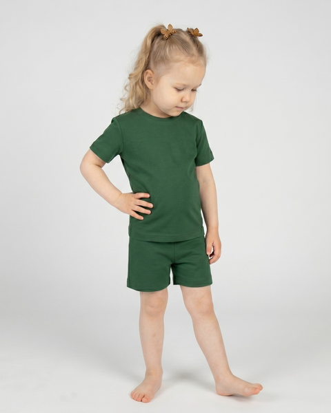 A young barefoot child stands against a plain light background wearing a dark green short-sleeve t-shirt and matching shorts. With one hand resting on their hip, they look down thoughtfully toward the floor, their blonde hair styled in half-up pigtails with small brown clips.