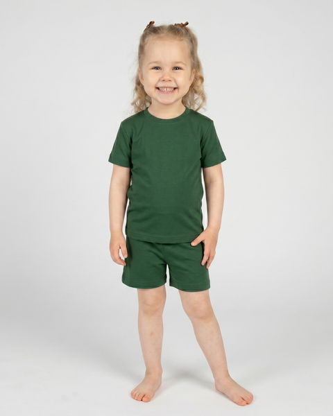 A smiling young child stands barefoot against a plain light background wearing a dark green short-sleeve t-shirt and matching shorts. They look directly at the camera with a bright, joyful expression, their hands resting gently near their sides and their hair styled in half-up pigtails.