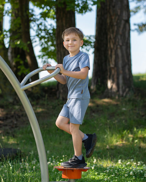 Young boy playing on outdoor playground equipment while wearing a dusty blue Merino wool T-shirt and matching shorts. The lightweight, breathable kids' outfit is showcased in a natural park setting, designed for comfortable movement during active play.