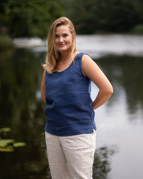 A woman stands near a calm river with her hands clasped comfortably behind her back, smiling softly at the camera. Her outfit consists of a dark blue sleeveless linen tank top paired with natural beige linen pants.