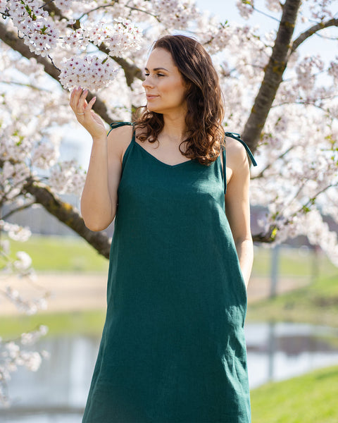 A woman with brown hair, wearing a long, dark green sleeveless dress with tie straps, stands outdoors next to a tree with white blossoms, touching the flowers.