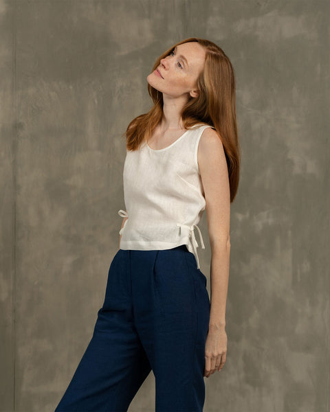 A studio shot of a woman with long, reddish-brown hair, looking upwards and to her left with a serene expression. She is wearing a white sleeveless linen top with side ties at the hem, paired with high-waisted, pleated blue trousers. Her arms are down at her sides. The background is a plain, textured grey wall.