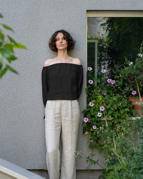 A woman with short, curly dark hair stands against a textured grey wall, looking directly at the camera. She is wearing a black off-the-shoulder top and light-colored linen trousers. To her right, a window is partially obscured by lush green plants with pink flowers in a terracotta pot.