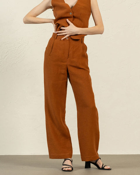 A person is shown from the mid-torso down, modeling an almond brown linen sleeveless vest and menique wide-legged trousers. One hand rests on their hip, while the other gently touches their stomach. They are wearing black open-toe heeled sandals. The background is a textured light beige wall and a light gray floor.