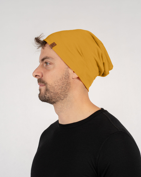 A man with short brown hair and a beard is shown in a profile view, wearing a spicy yellow merino wool beanie and a black t-shirt. He appears to be looking off to the left. The background is plain white.