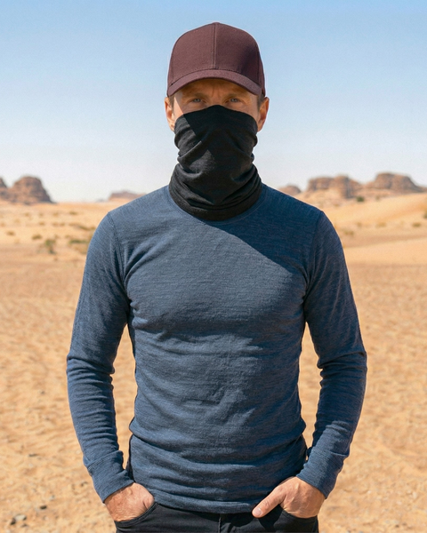 A man standing outdoors in a sandy desert landscape with rocky hills in the background under a clear blue sky. He is wearing a dark maroon baseball cap, a black neck gaiter pulled up to cover his nose and lower face, a textured blue long-sleeved shirt, and black pants. His hands are resting casually in his front pockets as he looks directly at the camera.