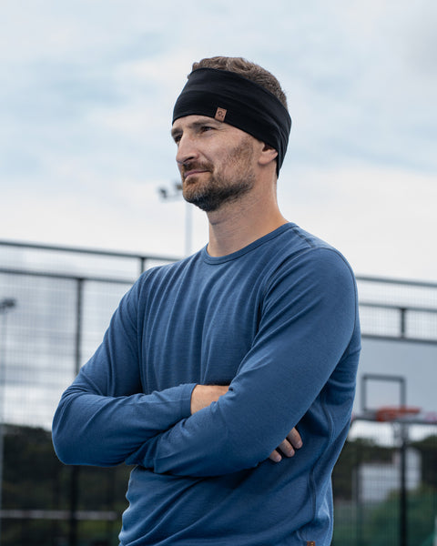 A man standing outdoors on a sports court with his arms crossed, looking off to the side with a neutral, focused expression. He is wearing a denim-blue long-sleeved shirt and a wide black headband. Behind him, a chainlink fence and a basketball hoop are visible under a cloudy sky.