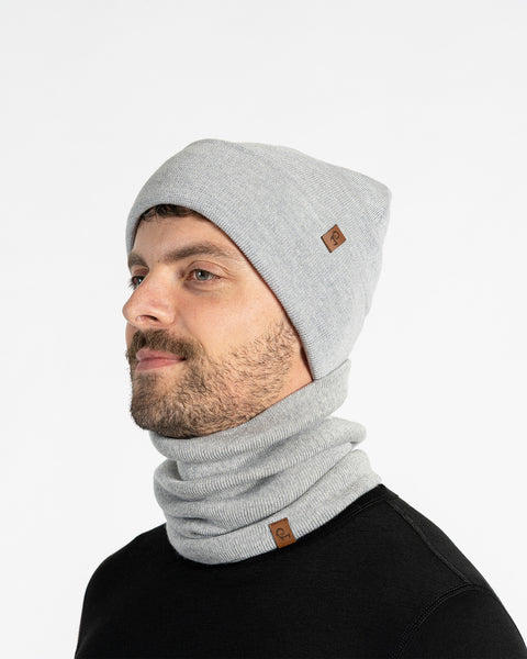 A man with a light brown beard is shown from the chest up, looking to the left of the frame. He is wearing a light gray knitted beanie and a matching neck gaiter. Both items of clothing have a small brown leather patch with a menique logo on them. The background is a plain white.
