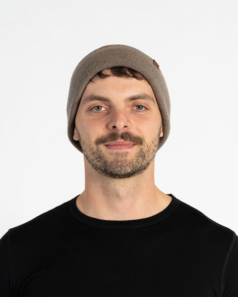 A person with light skin, short brown hair, a mustache, and light-colored eyes is shown in a headshot against a white background. They are wearing a brown knit beanie with a small brown tag on the side and a black long-sleeved shirt. They are looking directly at the camera.