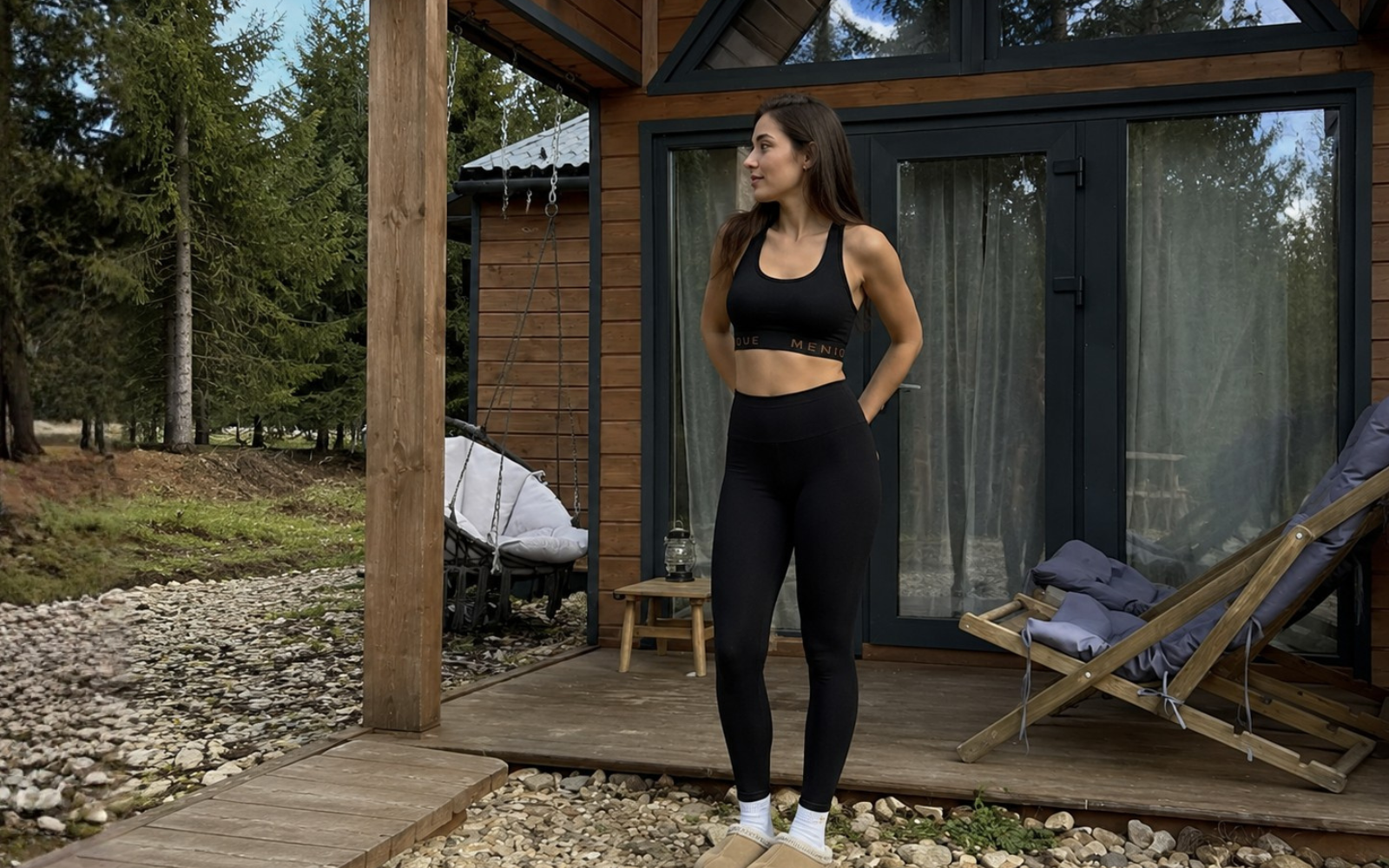 A woman wearing a black sports bra and black leggings standing on the wooden deck of a modern cabin surrounded by trees.