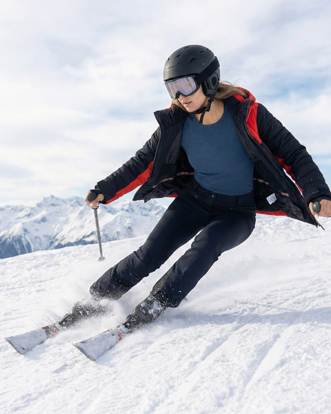 Woman skiing downhill on a snowy mountain wearing a denim blue merino wool base layer set under a ski jacket, with alpine scenery in the background.