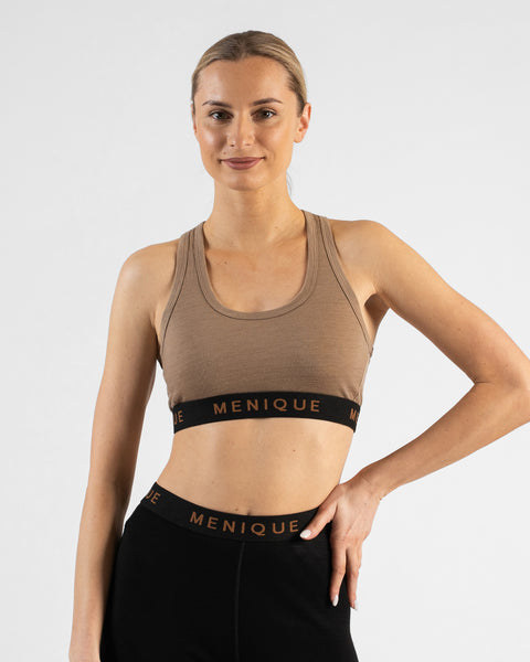 A studio portrait of a blonde woman smiling directly at the camera. She is wearing a beige sports bra with a black "MENIQUE" logo band and matching black leggings. She has one hand resting on her hip against a plain white background.