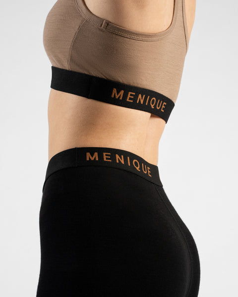 A close-up profile shot focuses on a person's midsection, highlighting a beige sports bra paired with black leggings against a plain light background. Both pieces of activewear feature a thick black elastic band displaying the brand name "MENIQUE" in bold copper lettering.