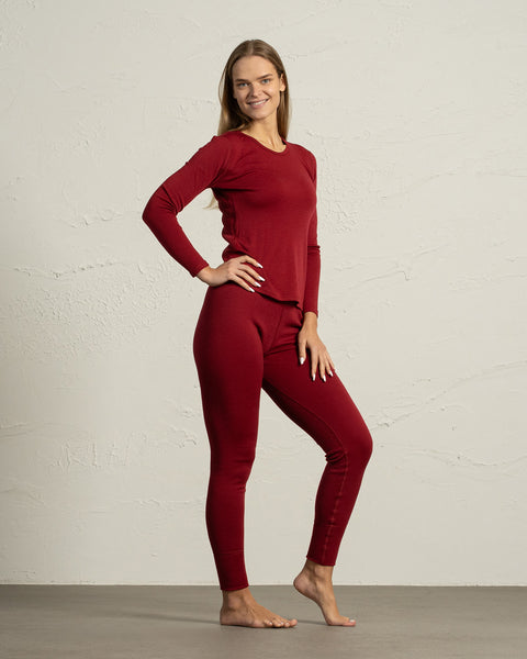 A full-body shot of a smiling model wearing a matching set of royal cherry red merino wool thermal underwear, standing barefoot with one hand on her hip.