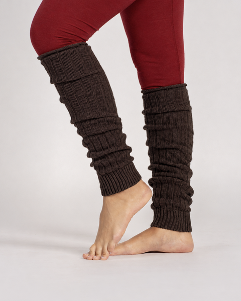 A view of a person standing barefoot, wearing brown, ribbed-knit leg warmers scrunched down over red leggings against a solid light gray background.