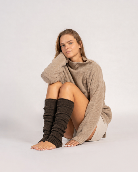 A view of a sitting woman barefoot, wearing brown, ribbed-knit leg warmers scrunched down against a solid light gray background.