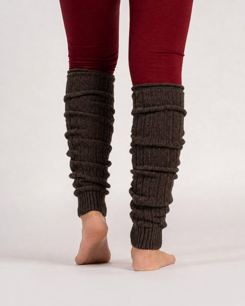 A person shown from the back wearing dark brown ribbed knit leg warmers over burgundy leggings, standing barefoot against a light gray studio background.