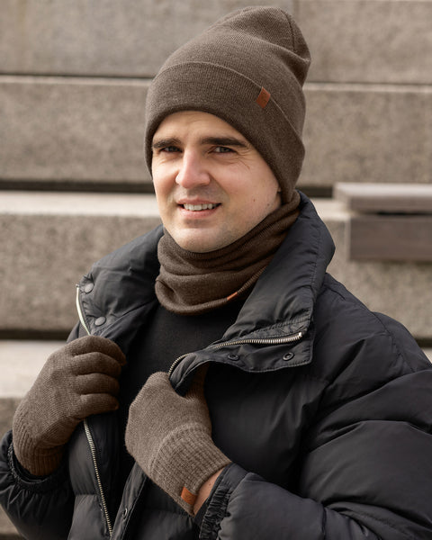 Man dressed in a brown Merino wool beanie, gloves, and neck warmer, paired with a black puffer jacket, posing on outdoor stone steps in winter.