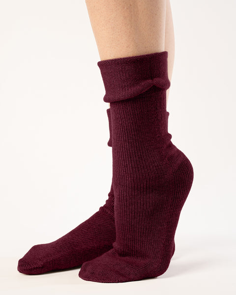 A side view captures a person from the mid-thigh down, showcasing ribbed, royal cherry socks with a partially folded cuff. The socks reach the mid-calf, and the person's feet are positioned with one slightly behind the other, heel raised. They stand against a stark white backdrop with even lighting that accentuates the texture and gathered folds of the socks.
