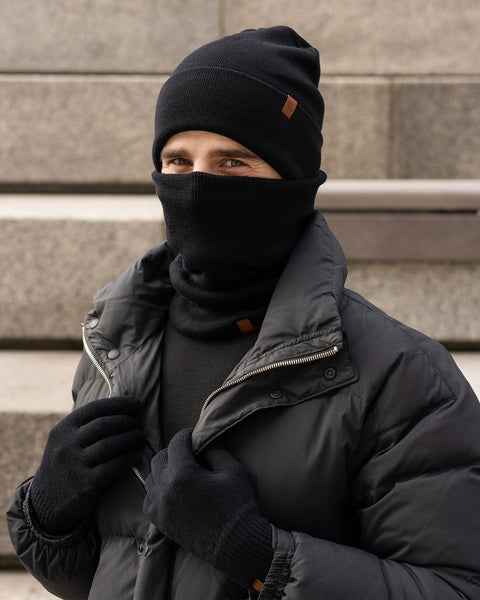 A person with short dark hair and stubble wears a black beanie, a black neck gaiter pulled up over their nose and mouth, a black puffer jacket, and black gloves. Only their eyes are visible. They are outdoors with concrete stairs in the background.