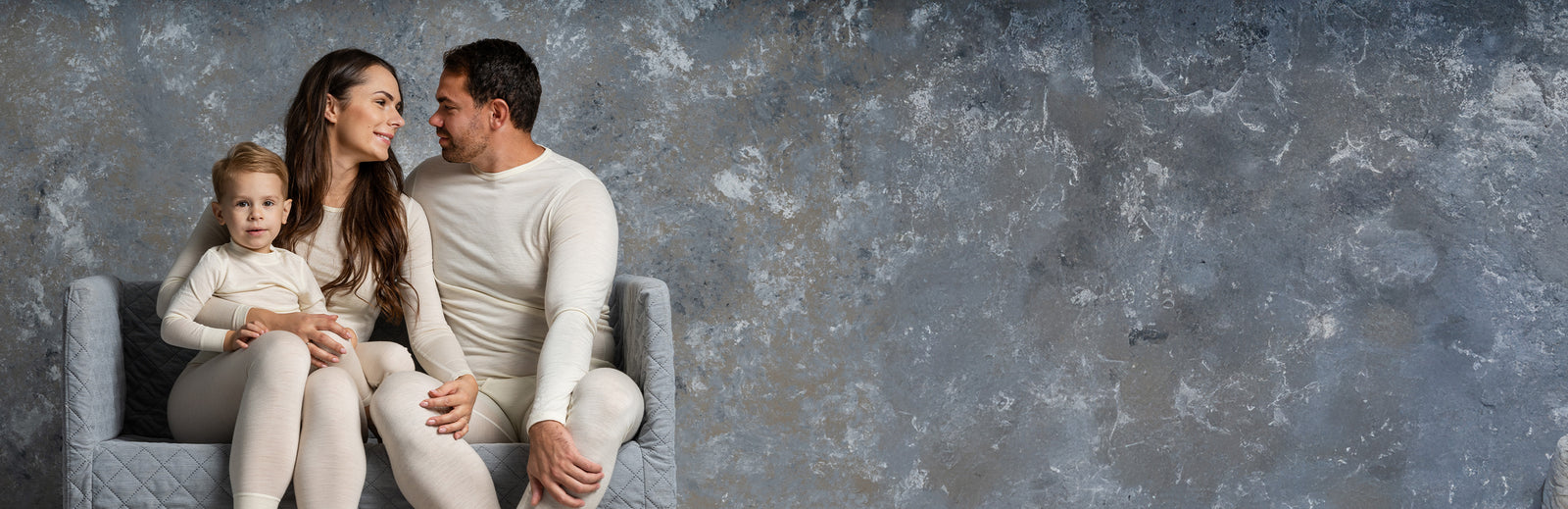 Family of three sitting together on a gray chair against a textured gray wall.