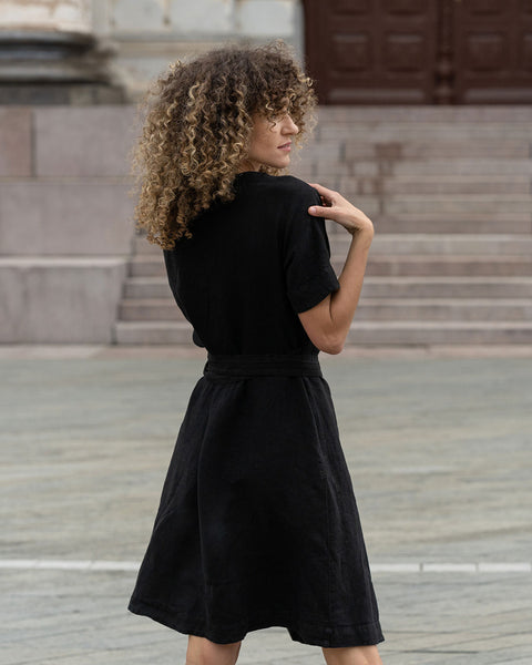 A beautiful woman with curly hair posing outside in the city from the back, wearing pure black linen dress Eliana