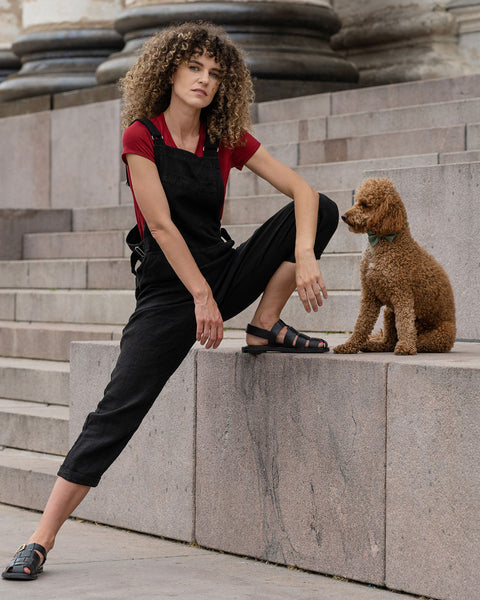 Beautiful woman with curly hair posing in the city wearing pure black linen jumpsuit Nicci, royal cherry merino wool top. Posing with a brown poodle dog.