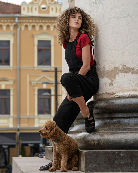 Beautiful woman with curly hair posing in the city wearing pure black linen jumpsuit Nicci, royal cherry merino wool top. Posing with a brown poodle dog.