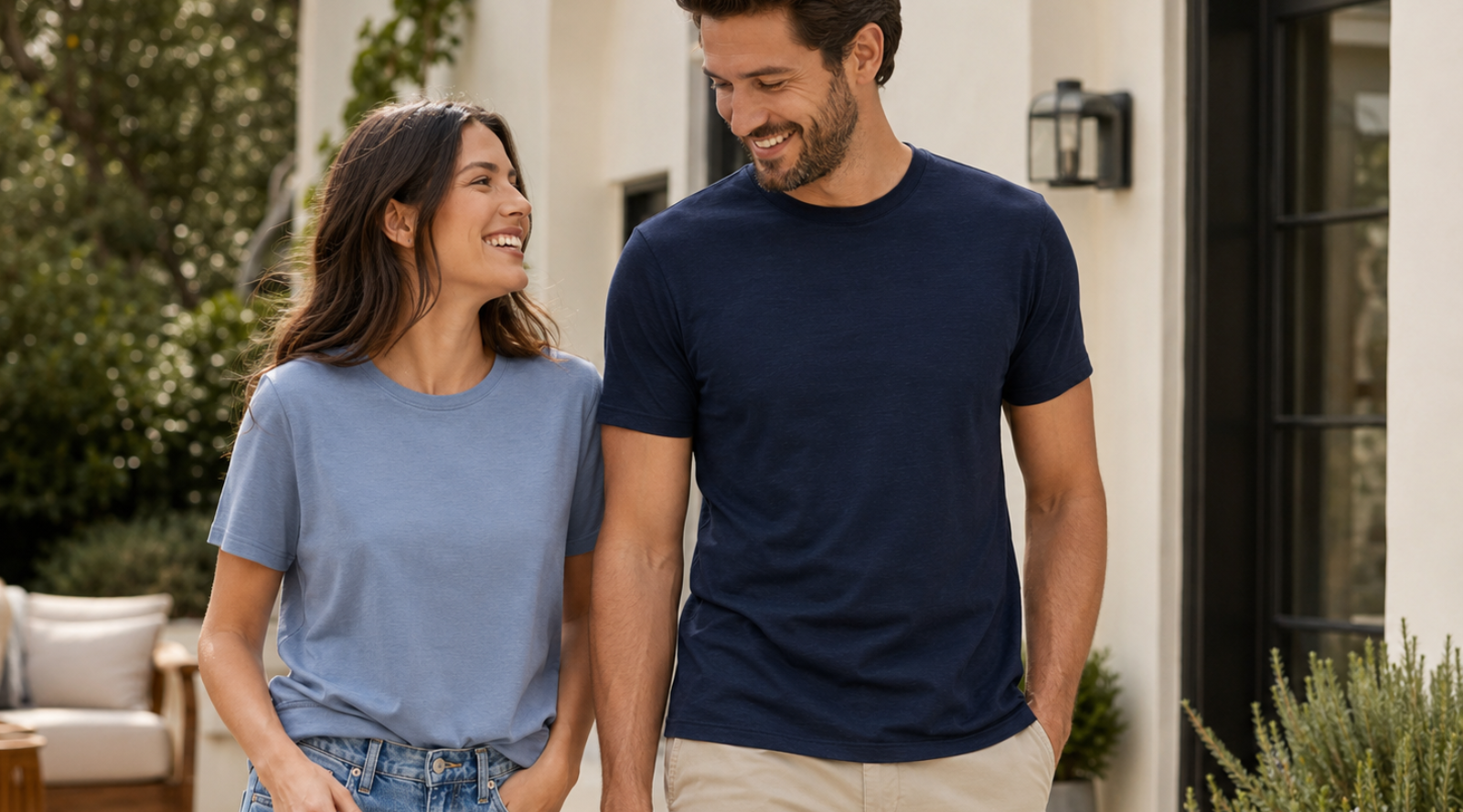 A smiling man and woman walking together outdoors near a white house. The woman is looking at the man and wearing a light blue short-sleeve t-shirt with blue jeans. 