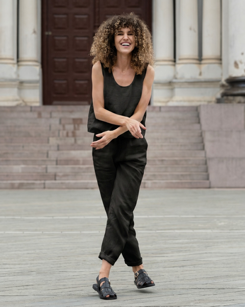 A woman with voluminous curly hair viewed from behind, wearing an olive green sleeveless linen top and matching pants. Her arms are slightly outstretched, and she is standing in front of wide stone steps leading up to dark wooden doors.