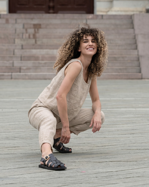 A smiling woman with curly hair crouching down in a stone plaza. She is wearing a light beige, natural-colored linen set with a sleeveless top and cropped pants, paired with black strappy sandals.