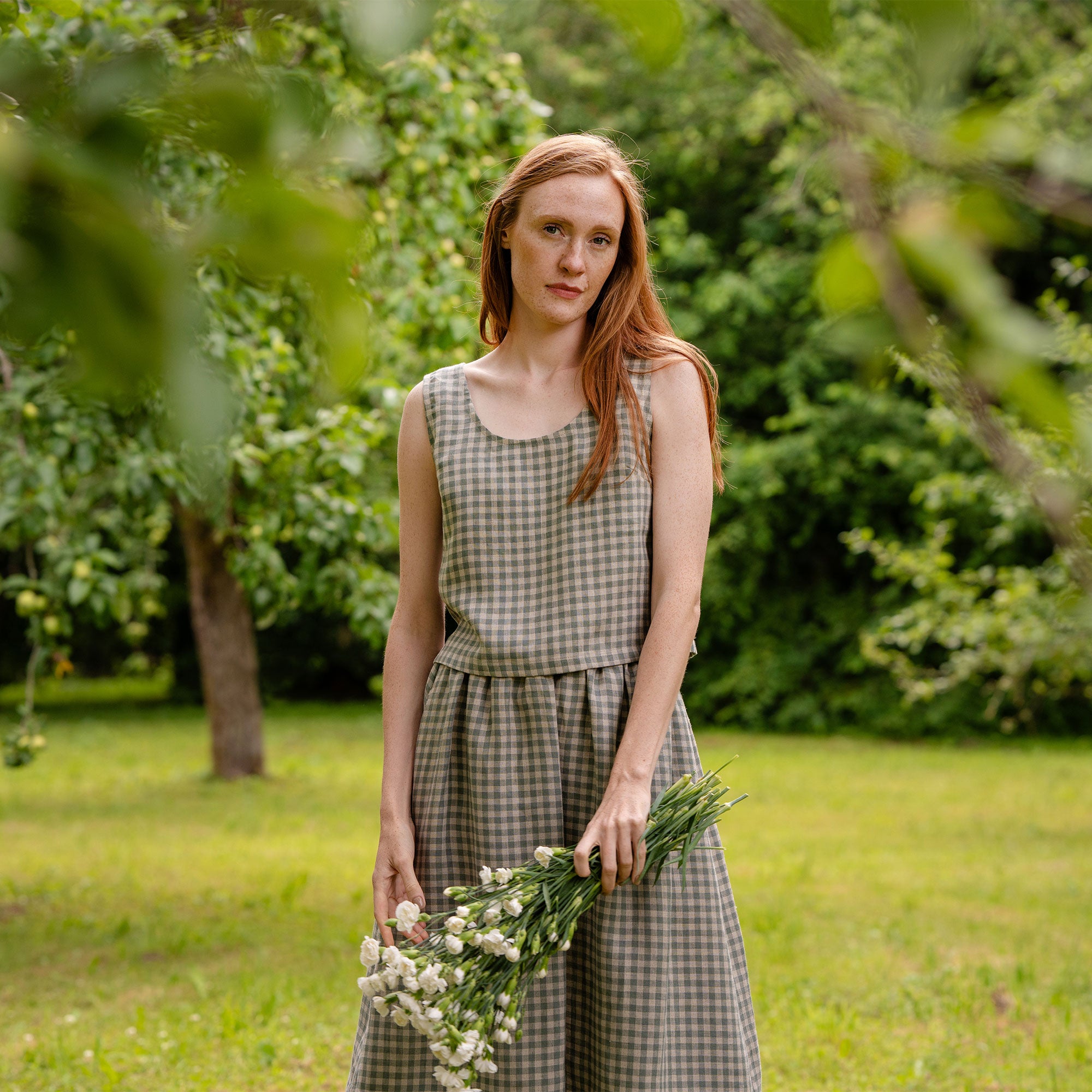The model in the photograph is visible from the front, with a close-up view of the ANA cropped linen blouse in a green gingham pattern. The top is sleeveless, matched with same garment bottoms. There are blurred nature elements visible in the background.