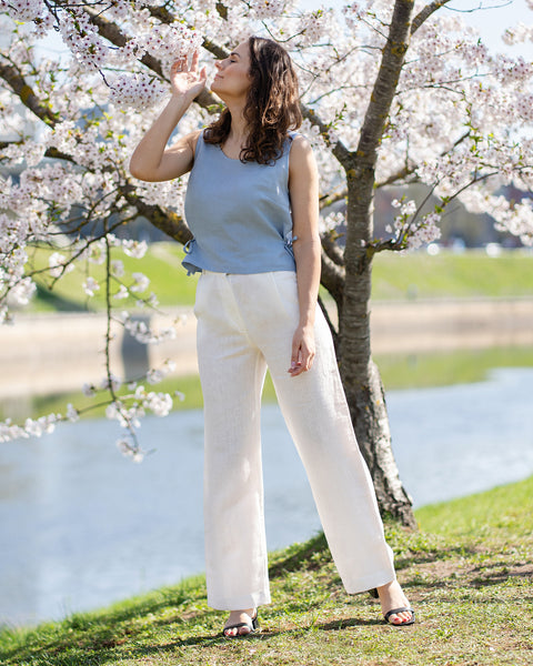 A woman in a cloudy blue sleeveless linen top with side ties and white wide-leg linen pants stands next to a blooming cherry blossom tree. She has her hand near her face as if smelling the blossoms, with a serene body of water and green grass in the background.