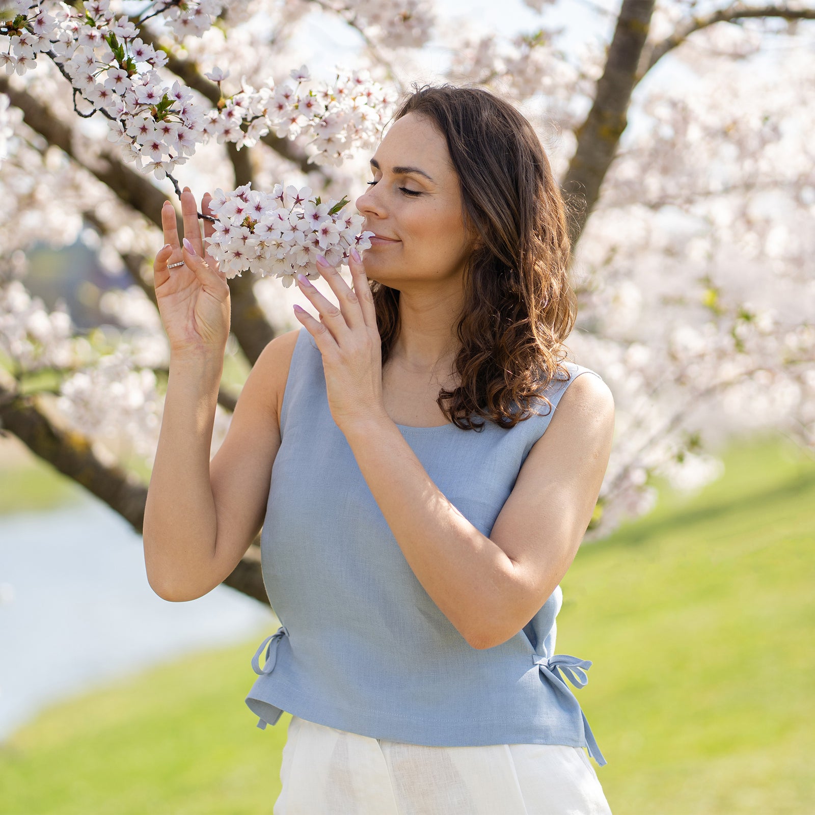 A smiling woman with dark, wavy hair wears a cloudy blue sleeveless linen top with side ties, paired with white linen pants. She stands outdoors under a blooming cherry blossom tree, with a body of water and green landscape in the soft background.