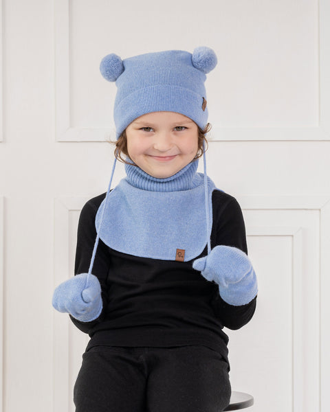 A young child with light brown curly hair and a subtle smile, is looking directly at the viewer. They are wearing a light blue knit hat with two pom-poms and earflaps that tie under the chin, matching light blue mittens, and a light blue knit neck warmer, over a black long-sleeved shirt. The background is a white paneled wall.