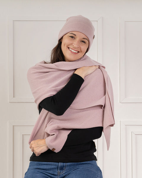 A smiling woman looking directly at the camera. She is wearing a dusty pink knit beanie and a matching dusty pink knit wrap gracefully draped over her shoulders. She is dressed in a black long-sleeved top and blue jeans, posed against a white paneled wall.