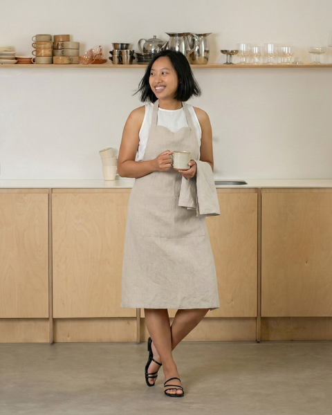 Woman wearing a natural linen bib apron, standing in a modern kitchen and holding a ceramic mug.