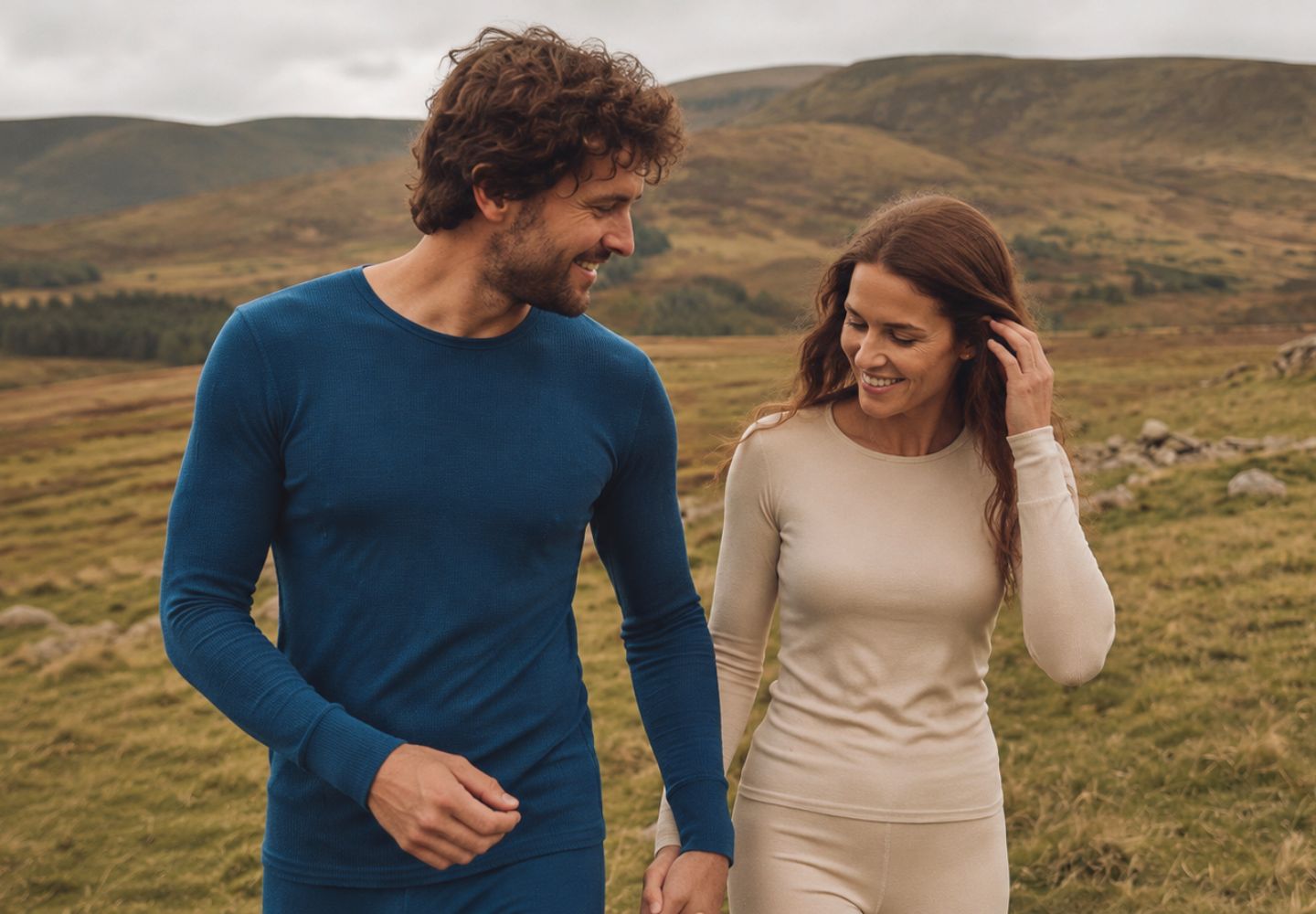 A man and a woman walking hand in hand through a grassy mountain landscape, wearing matching long-sleeve knit base layers in deep blue and light beige.