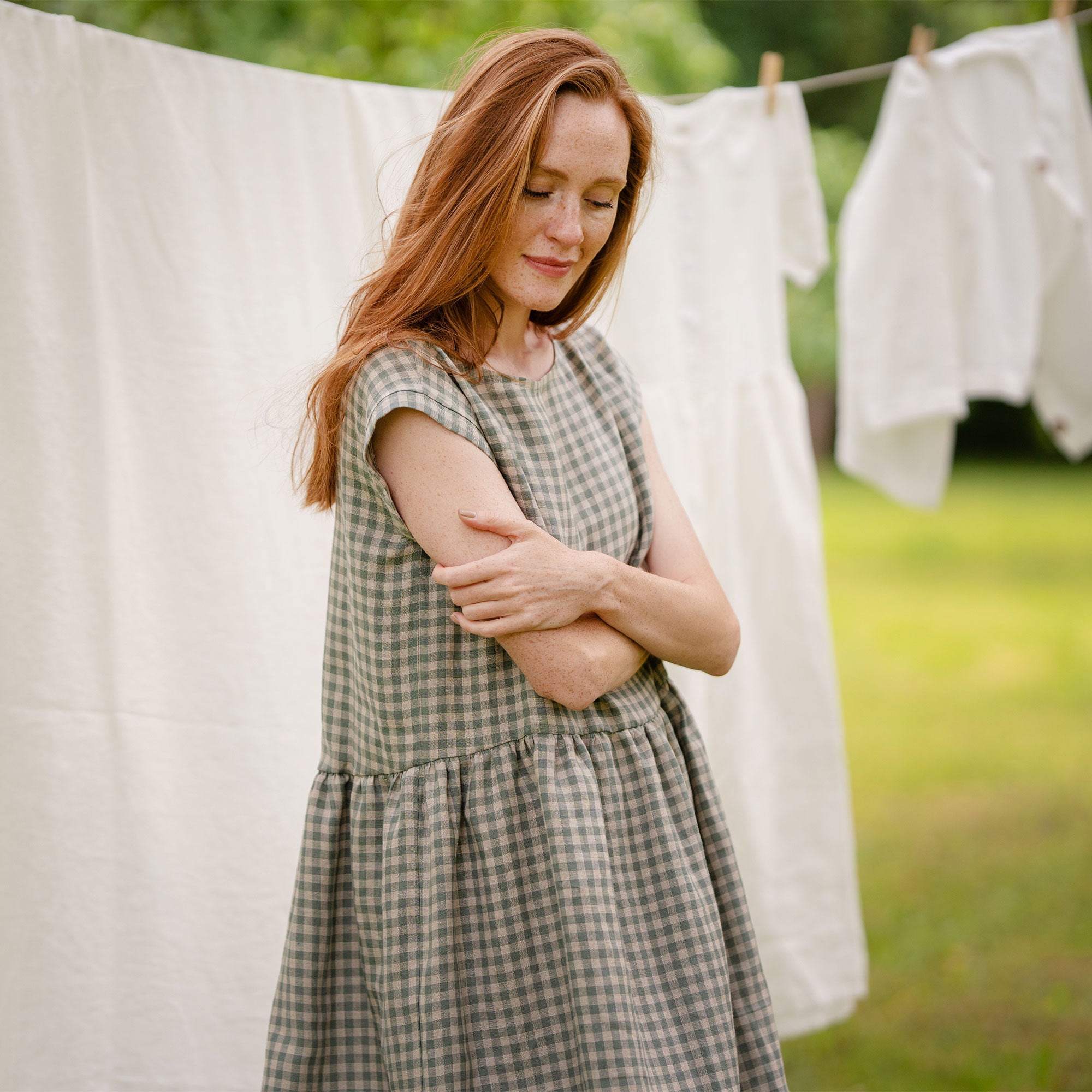 The model is wearing a cap sleeve, knee-length gingham dress in a light green and white checkered pattern, standing barefoot outdoors near a clothesline with white garments hanging in a lush green garden.