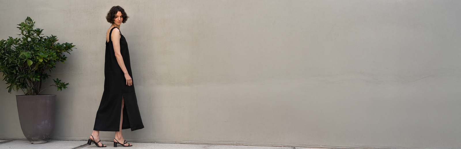 A woman wearing a black sleeveless linen dress with a side slit, photographed in profile against a light neutral wall. The minimalist summer look highlights the natural texture of linen and the relaxed, flowing silhouette of the dress.