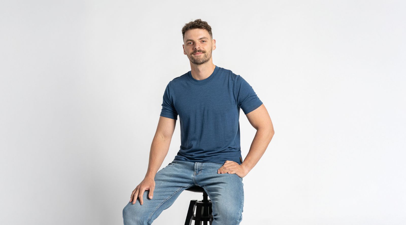 Man sitting on a stool wearing a blue t-shirt and jeans against a light gray background
