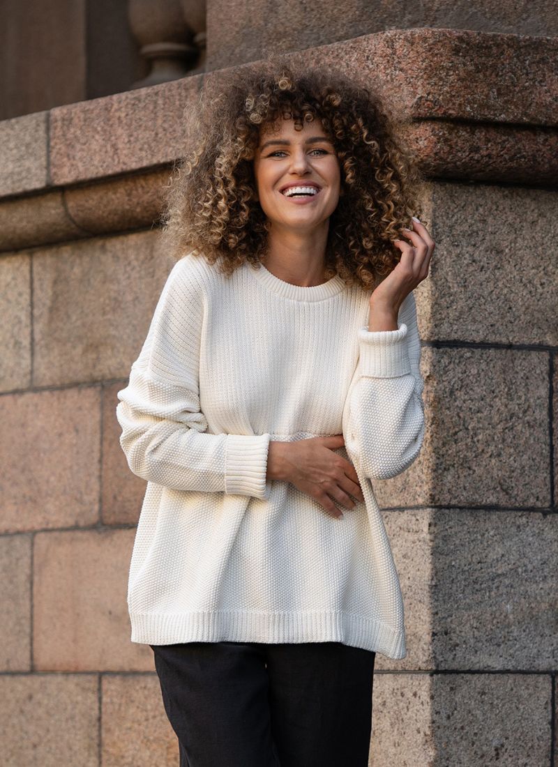 Woman with curly hair smiling and wearig white knit sweater.